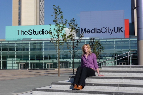 Angela Stevenson, wearing a purple top with black trousers, sitting on the steps outside the main TV studio block at MediaCity UK
