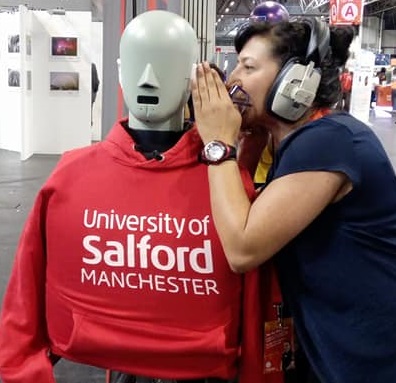 Philippa wearing headphones and shouting into the ear of a grey mannequin wearing a red University of Salford Manchester t-shirt