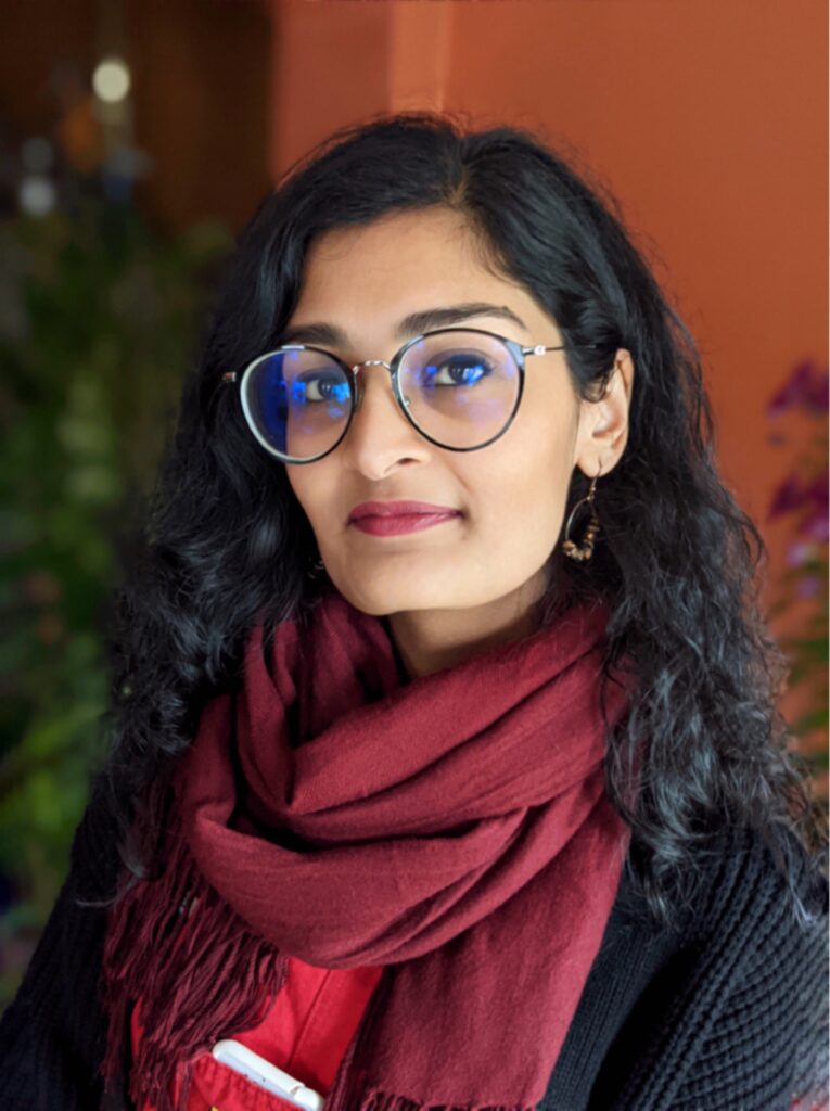 Regina Chandran - South Asian woman with long black hair and glasses, wearing a burgandy red scarf and looking at the camera.