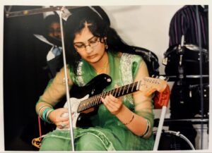 Regina Chandrand - Asian woman with long, dark hair and glasses wearing a spring green shalwar kameeze as she sits to play her electric guitar.