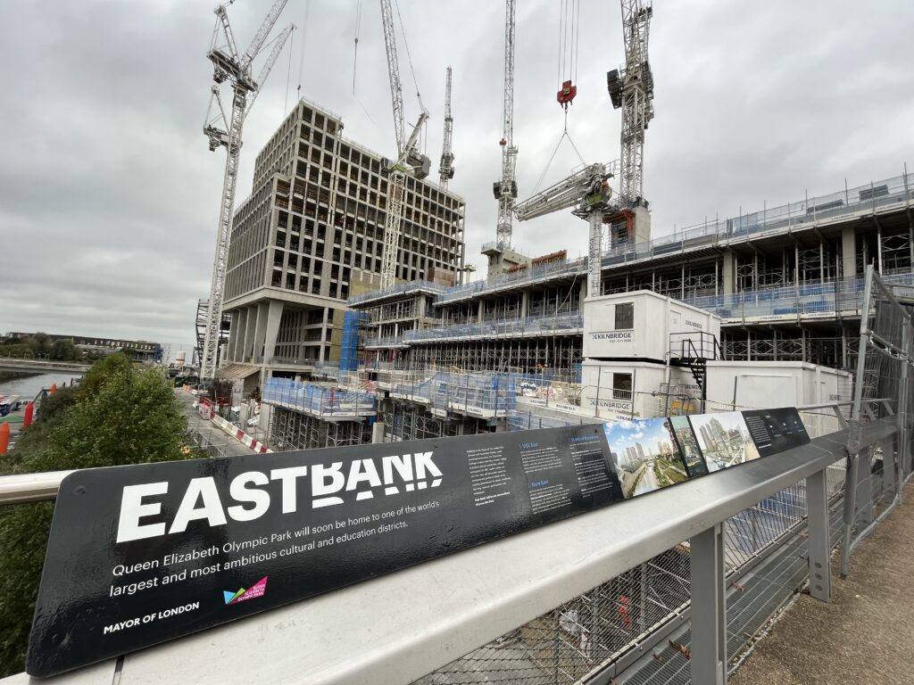 A concrete skeleton of a building being constructed by cranes with a black and white East Bank sign in front. Photo credit: BBC.