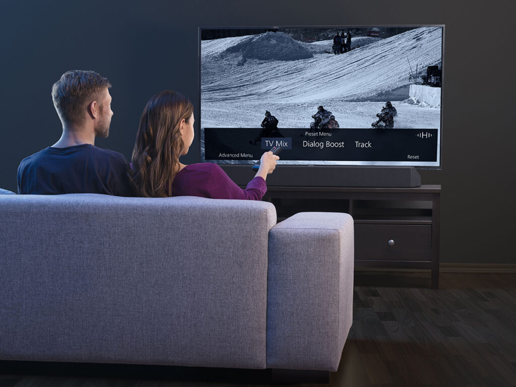 A man and a woman face away from us as they sit on the sofa and point their remote at a large television. Credit: Fraunhofer