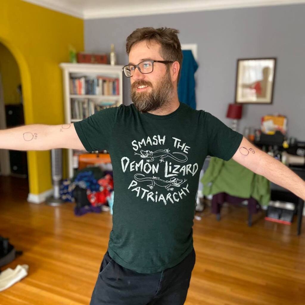 Martin Austwick - White man with beard and glasses wearing a bottle green t-shirt which says 'smash the demon lizard patriarchy'.