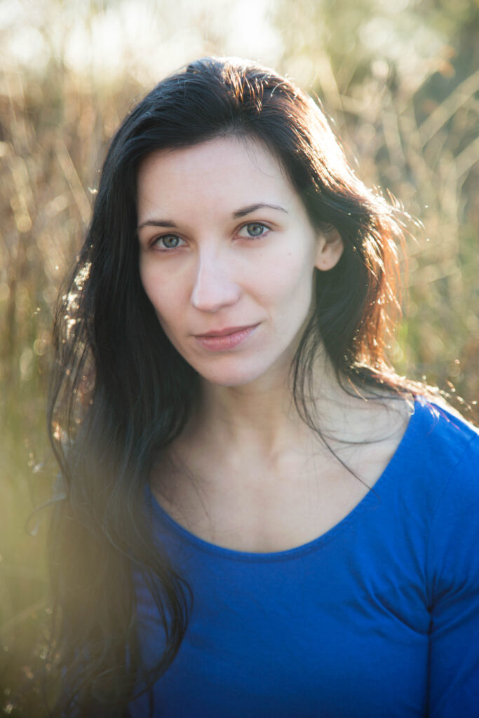 Ioana Barbu - White woman with very long dark brown hair and blue eyes wearing a royal blue scoop-neck t-shirt in a cornfield.