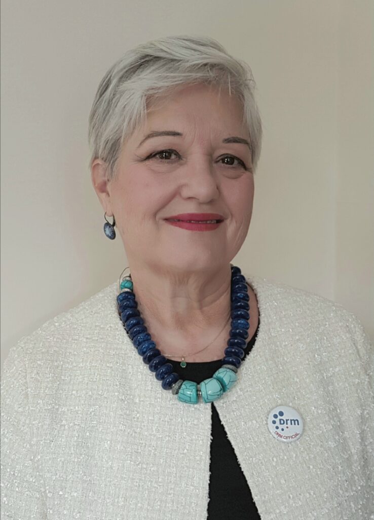 Ruxandra Obreja - White older woman with dark eyes, light grey hair, pink lipstick and wearing a white jacket with a turquoise three bead necklace.