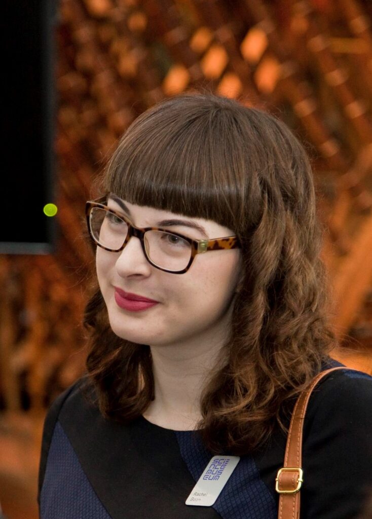 Rachel Boon - White woman with curly shoulder length hair and a fringe wearing brown tortoiseshell glasses and looking away from the camera.