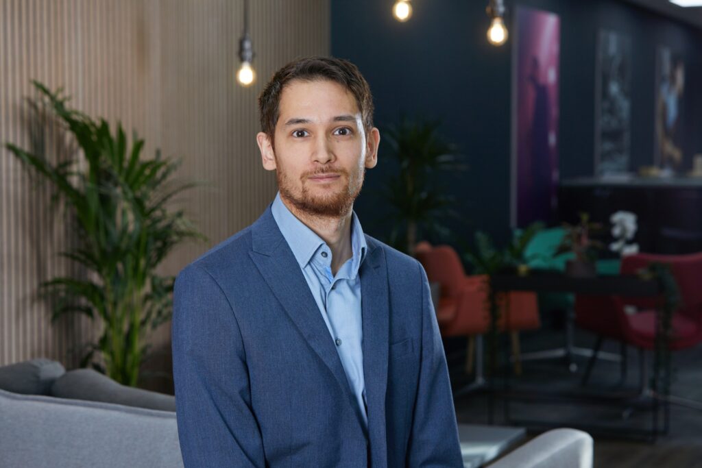 Andy Land - Man with brown hair and beard and dark brown eyes wearing a blue suit with a blue shirt. A trendy office with low-hanging lightbulbs is blurred in the background.