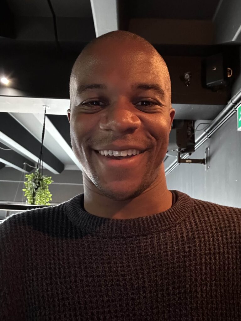 Thadeous Matthews - Man with brown skin and brown eyes wearing a brown fisherman style jumper. He's smiling at the camera and we can look up into the ceiling and lighting infrastructure above his head.