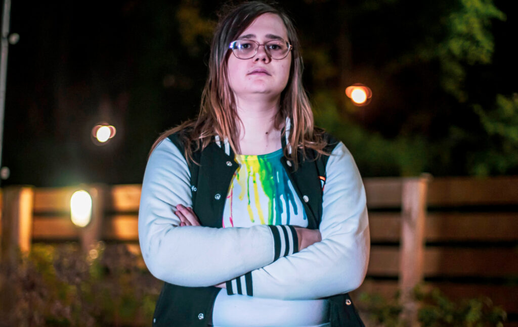Jamie Woods - white woman with glasses and long brown hair going down past her shoulders. She's wearing a grey top with rainbow streaks from the top that look like paint has been dribbling downwards in an artful fashion. Over the top is a black open waistcoat. She's looking down slightly towards the camera. Her arms are folded and the blurred streetlights in and fence in the background give the shot a dramatic feel.