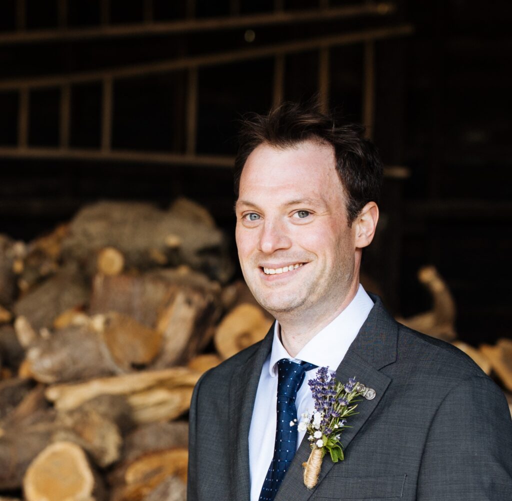 Mark Farrington - white man with short brown hair smiling in a wedding suit with lavender buttonhole.
