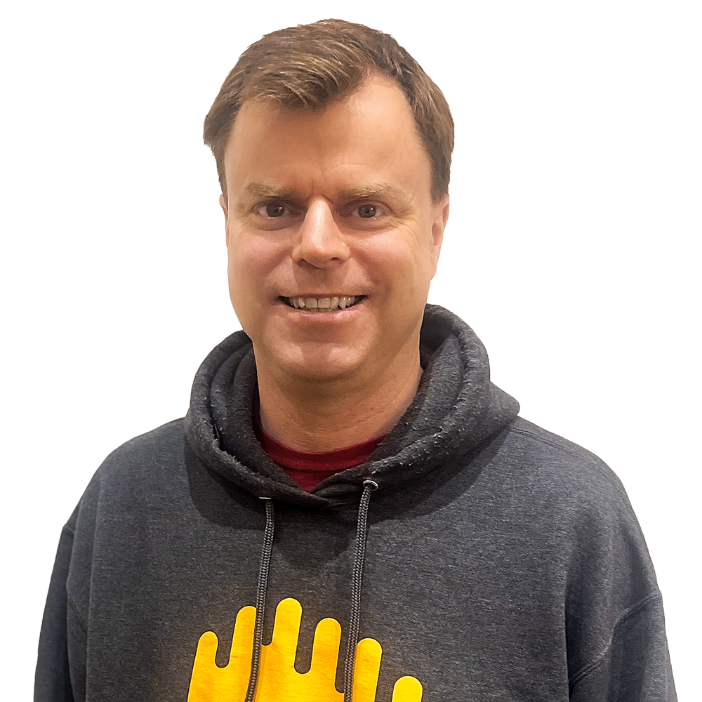 Dan - white middle aged man with short brown hair grinning at the camera. He's wearing a comfy grey fleece with a yellow-orange Broadcast Bionics sound/brain logo.