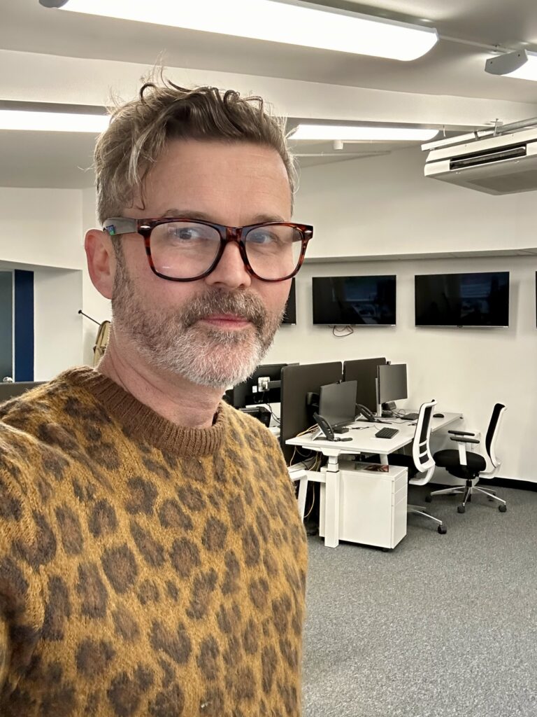 James - a man in his late forties with short beard and brown/graying hair - is looking straight at us with a friendly expression. He's wearing brown glasses and a leopard-print jumper. Empty office desk and chairs are in the background.