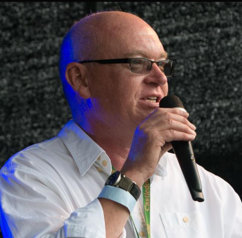 John - a white man with a bald head - is wearing tinted glasses and looking slightly to one side. He's holding a microphone close to his mouth and appears to be in the middle of hosting a live event.