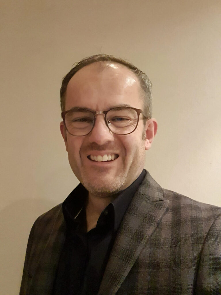 James - a white man with very short brown hair and even shorter and lighter beard - is wearing glasses and grinning at the camera. He's sporting a checked dark brown jacket with black shirt. 