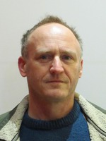 A slightly blurry ID card photo of Matthew - a white middle-aged man with blue eyes, short grey hair and wearing a sheepskin-style fleece jacked over a mid blue jumper.