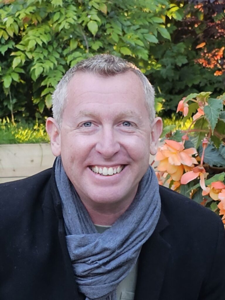 Richard - white man with light grey hair and blue/grey eyes wearing a light blue scarf and black overcoat smiling at the camera. He's in a garden with autumnal leaves behind.