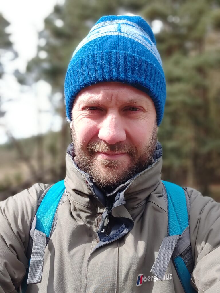 Richard - white man with light brown bear is squinting slightly in the camera as it's early morning and he's off on a hike, wearing a blue bobble hat and blue rucksack. 