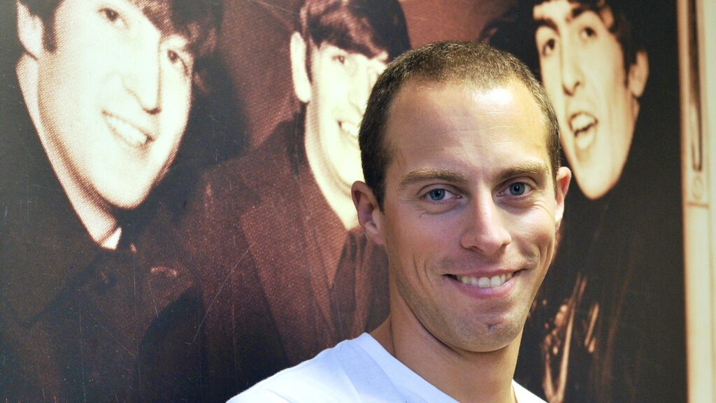 Simone - white man with short hair and blue eyes - is smiling at us. He's wearing a white t-shirt and standing in front of a large black and white picture of The Beatles.