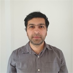 Daniyal - a man with light brown skin and dark hair with a very short beard and brown eyes - is looking at us and wearing a grey shirt with white buttons. The photo is a little blurry.